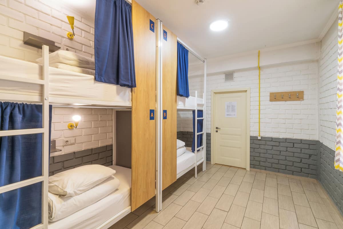 A dormitory room with bunk beds, a white brick wall, a wooden door, and a coat rack.