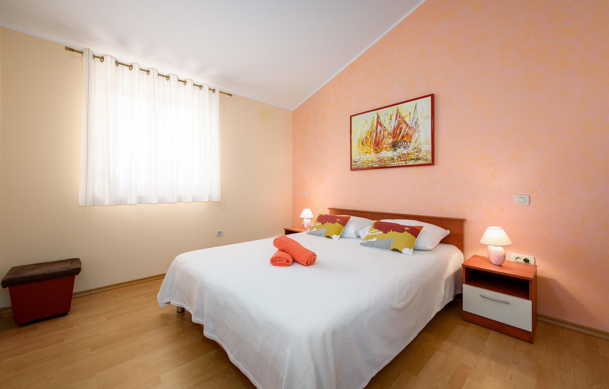 A neatly made bed with white linens and orange towels, a wooden nightstand with a lamp, and a painting on the wall.