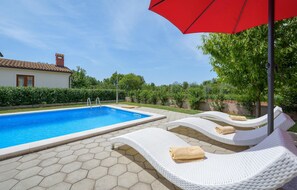 Ein Swimmingpool mit drei weißen Liegestühlen, jeweils mit einem Handtuch, und einem roten Sonnenschirm.