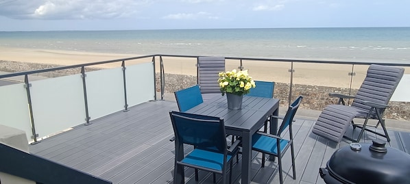 A deck with a table and chairs, a grill, and a view of the beach.