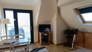 Una stanza con TV su un supporto in legno, un treppiede, un tavolo da pranzo con sedie e vista mare attraverso un'ampia finestra.