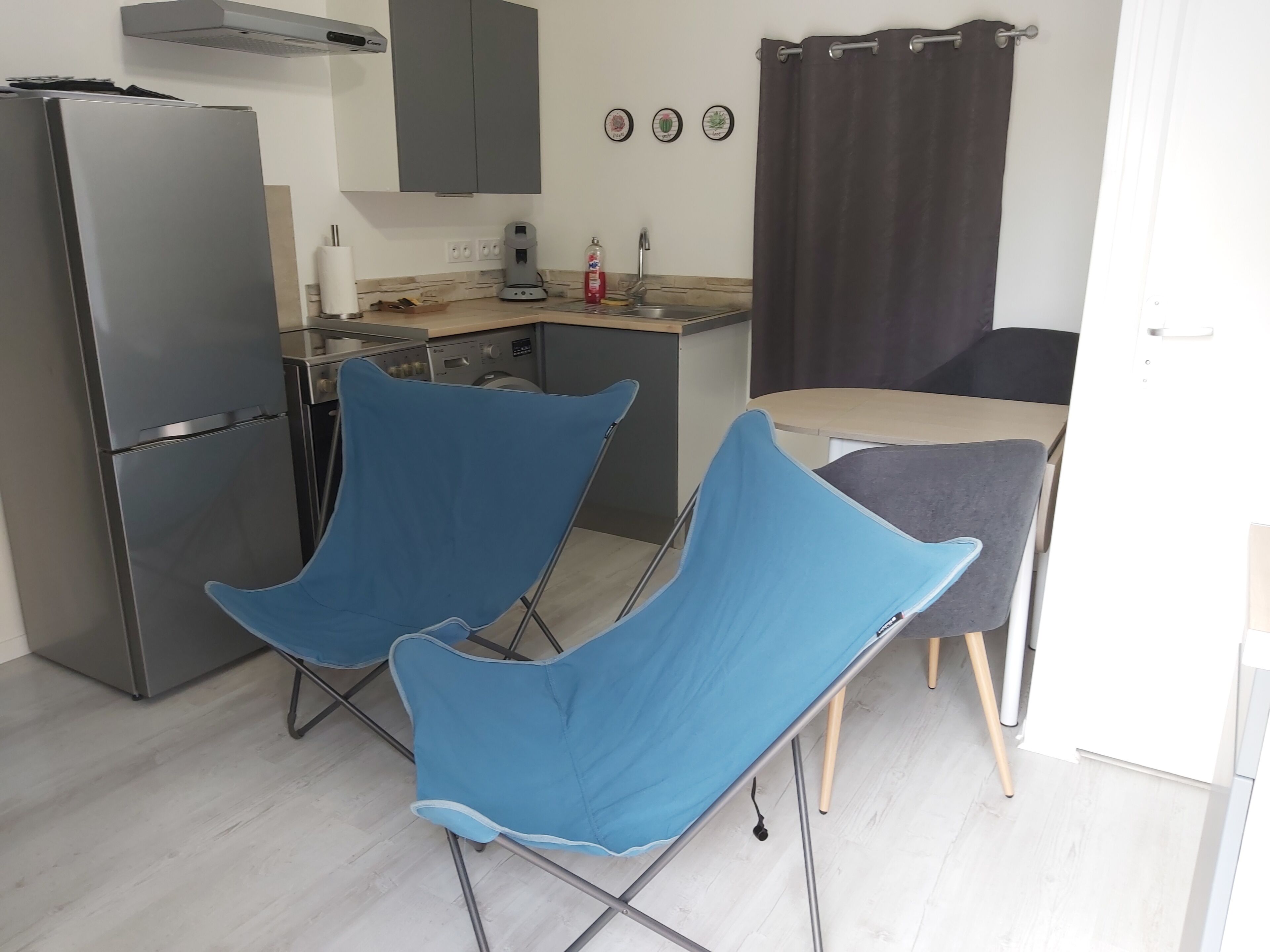 Une cuisine compacte équipée d’un réfrigérateur, d’une machine à café et d’un évier. On y trouve deux chaises bleues et une table avec un coussin gris.