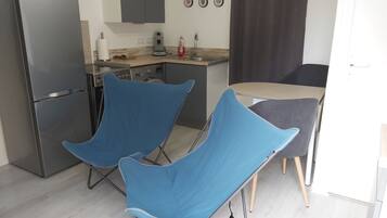 Une cuisine compacte équipée d’un réfrigérateur, d’une machine à café et d’un évier. On y trouve deux chaises bleues et une table avec un coussin gris.