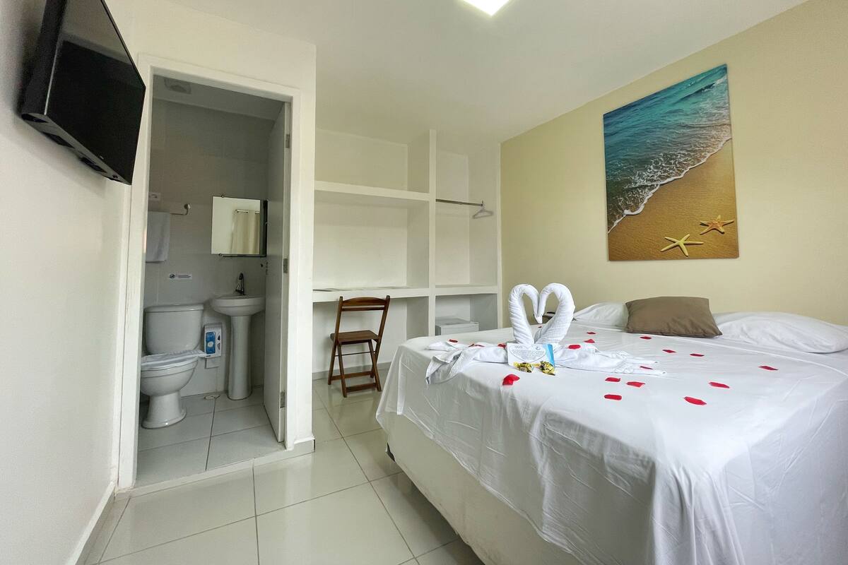 A hotel room with a bed, a painting of a beach, a television, and a bathroom visible through an open door.
