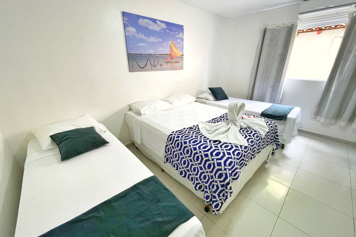 A hotel room with two single beds, a large bed with a patterned blanket, a painting of a sailboat, and a window with curtains.