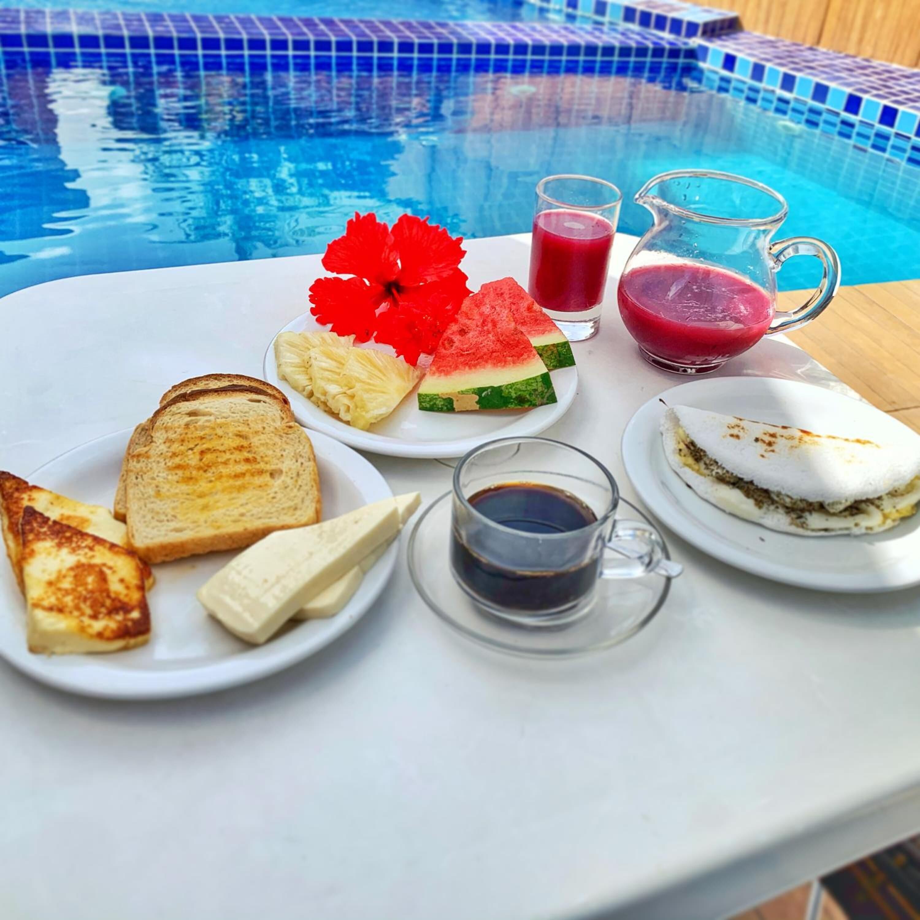 Un petit-déjeuner est préparé au bord de la piscine : il comprend des toasts, du fromage, une salade de fruits, un smoothie et une tasse de café.
