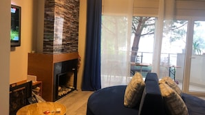 A living room with a stone fireplace, a round wooden table, a dark blue sofa with patterned cushions, and a large window with a view of trees and a balcony.