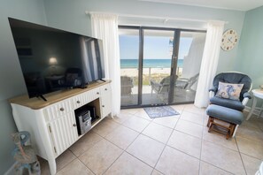 A living room with a flat-screen TV, a wooden entertainment unit, a beach view through a sliding glass door, and a blue armchair.