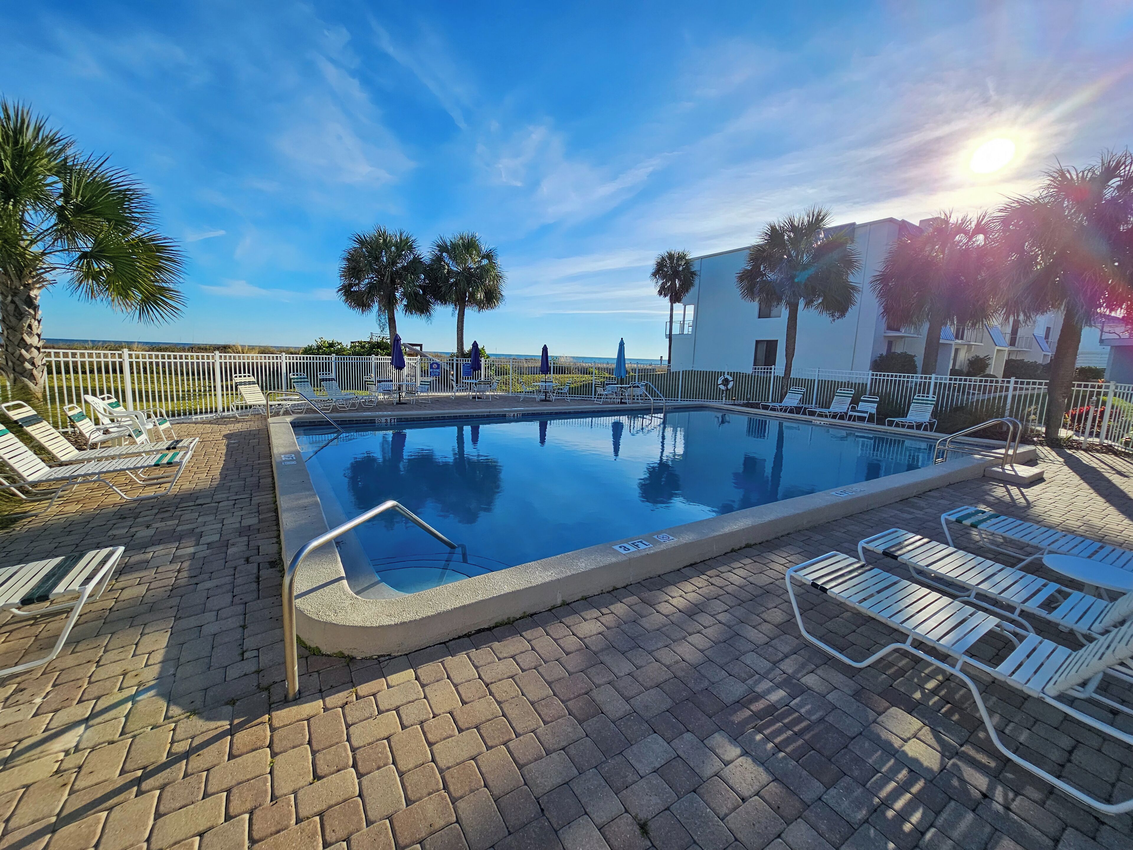 A swimming pool with lounge chairs and umbrellas, surrounded by a white fence and palm trees.