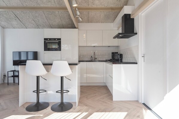 A modern kitchen with white cabinets, a black stove, and two bar stools. - Therese: Charming garden cottage. (Zandvoort)