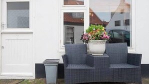 En hvid facade med en dør og vinduer. Der er en grå fletsofa med en hvid plantekasse med blomster.