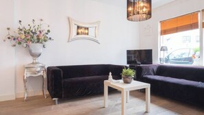 En moderne stue med en mørk fløjlssofa, et hvidt sofabord, en dekorativ vase med blomster, et spejl på væggen og en hængende lampe.