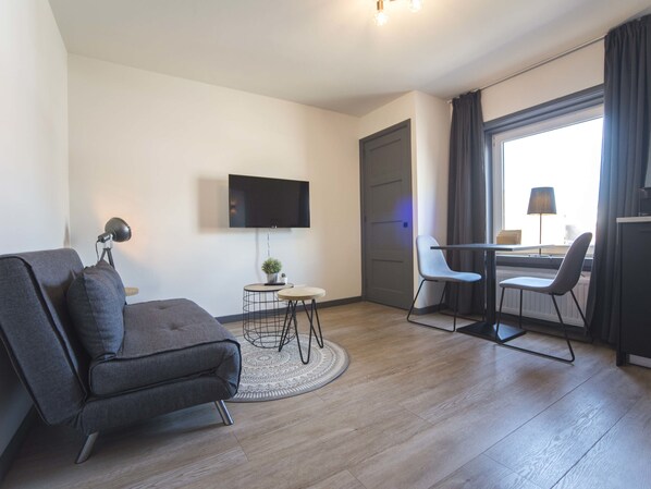 A modern living room with a sofa, a small round table with a plant, a TV mounted on the wall, and a dining area with a chair and table.