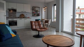 A modern kitchen with white cabinets, a built-in oven, and a stone backsplash. There is a dining area with a round wooden table and chairs, and a large window offering a view of the city. - De Boulevard A with private parking (Zandvoort)