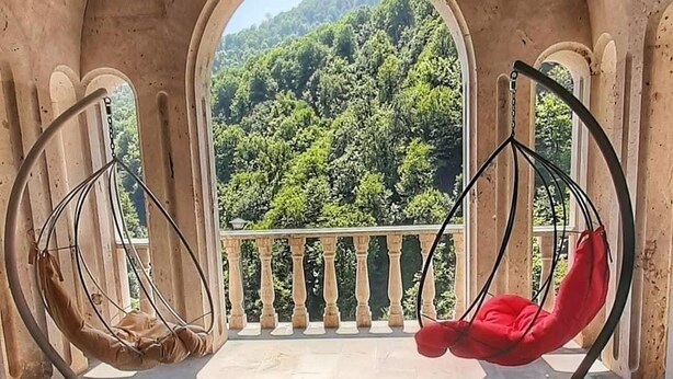 A balcony with two swing chairs, one beige and one red, overlooking a forested hillside. - Vanatun Monastery Stay (Dilijan)