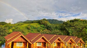 A row of wooden cabins with pitched roofs, set against a backdrop of lush green hills and a rainbow. - Phusiatfha Resort (Khao Kho)