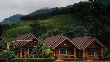 A row of wooden cabins with pitched roofs, surrounded by lush greenery and hills in the background.