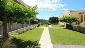 Un patio con un sendero pavimentado, rodeado de céspedes y setos cuidados, que conduce a un edificio de fachada color crema.