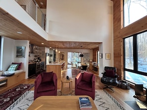 A spacious living area with a high ceiling, wooden beams, a dining table, two red armchairs, and a view through large windows.
