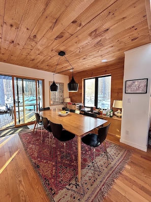 A wooden interior with a dining table, chairs, a rug, and large windows.