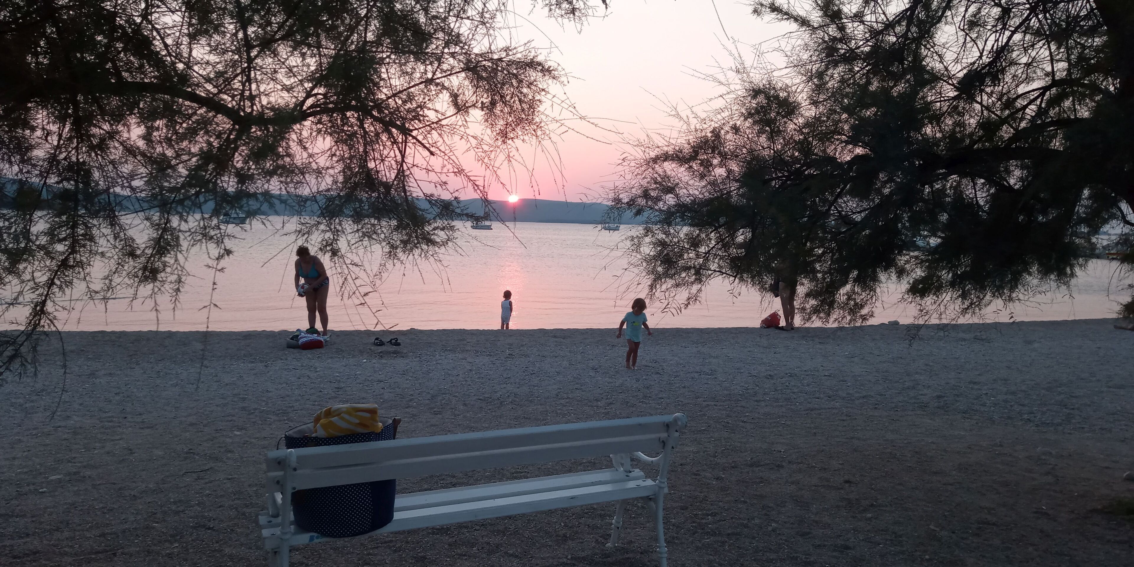 A bench, a child, and a person standing by a lake at sunset.
