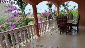 Un balcon doté d’une balustrade blanche, une table avec des chaises, et une vue sur des fleurs violettes et des bâtiments en arrière-plan.