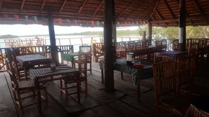 An outdoor dining area with wooden tables and chairs, a thatched roof, and a view of a lake. - Bintang Bolong Lodge (Kassagne)