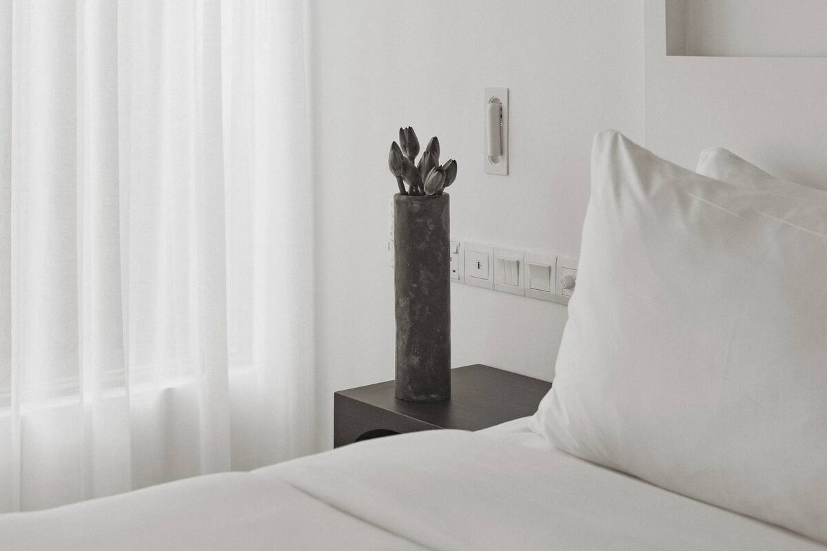 A minimalist hotel room with a large bed, a bedside table with a vase, and sheer curtains.