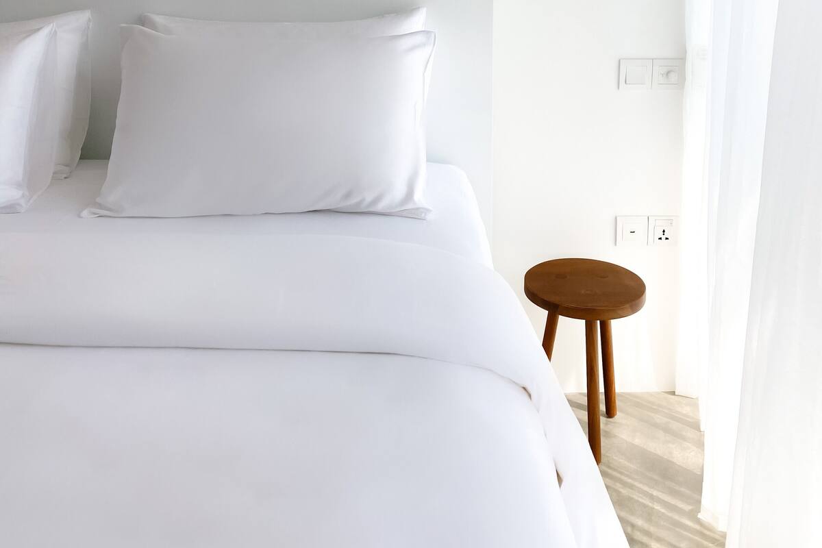 A minimalist hotel room with a large bed, a small wooden stool, and a plain white wall.