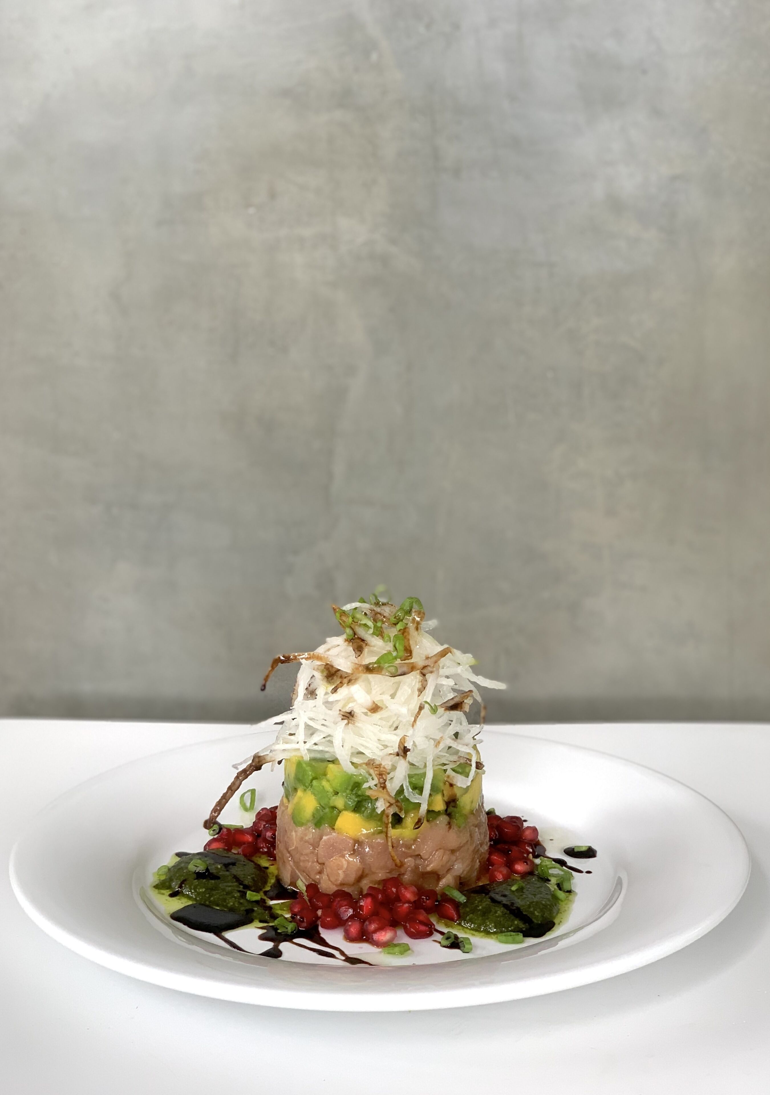 Un plat blanc amb un plat de menjar, decorat amb herbes verdes i llavors de taronger, davant d'un fons gris.