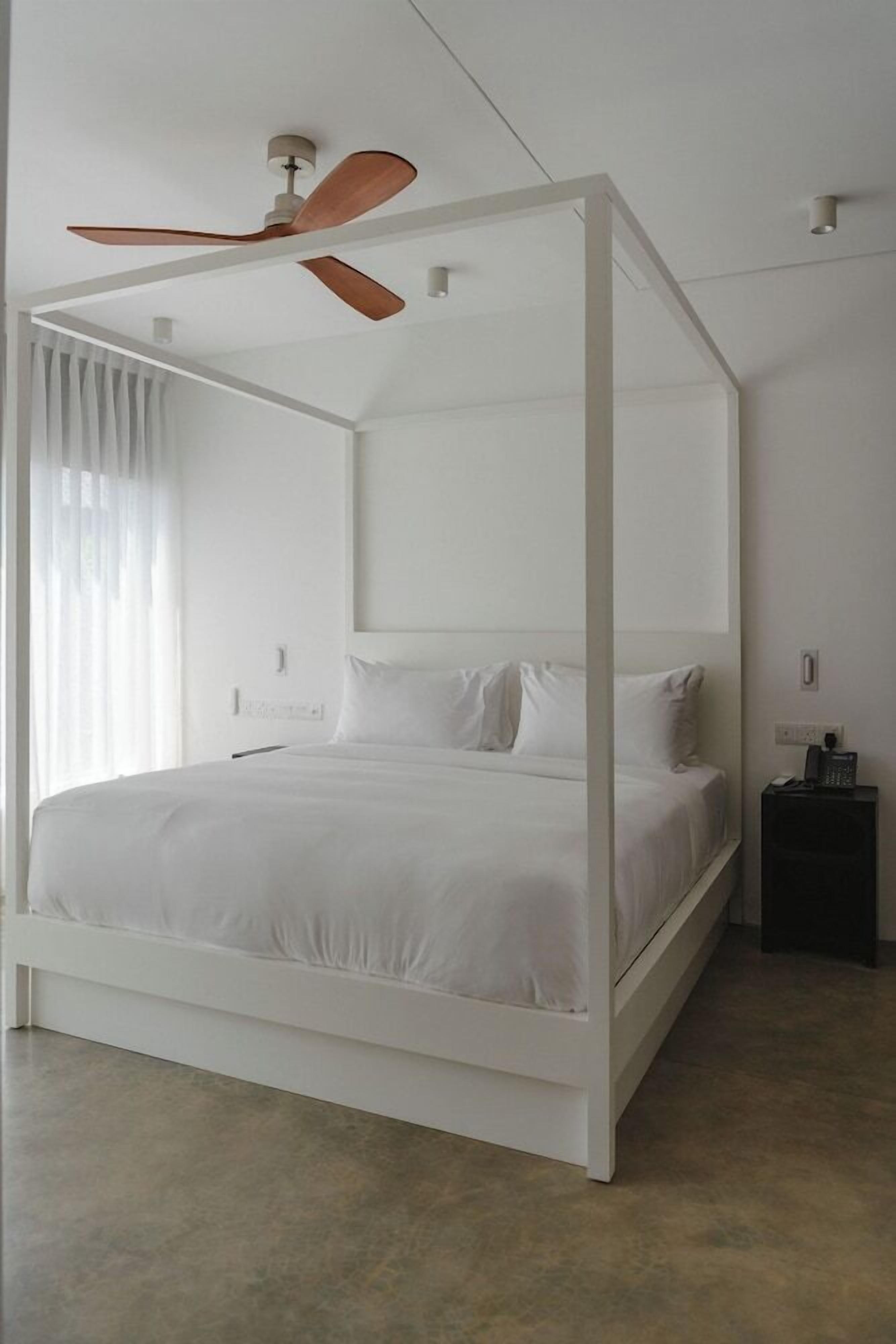 Uma cama de dossel branca com um ventilador de teto, uma mesa de cabeceira e uma janela com cortinas.