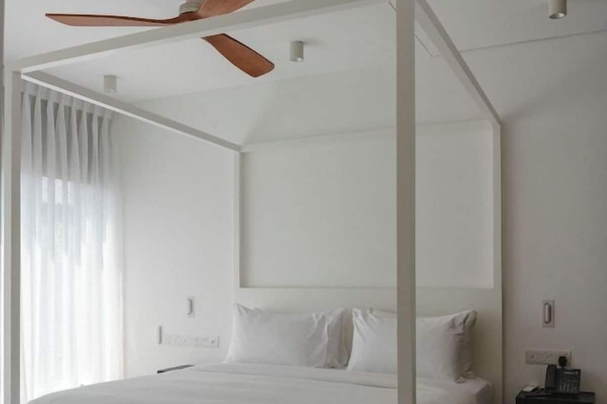 Uma cama de dossel branca com um ventilador de teto, uma mesa de cabeceira e uma janela com cortinas.