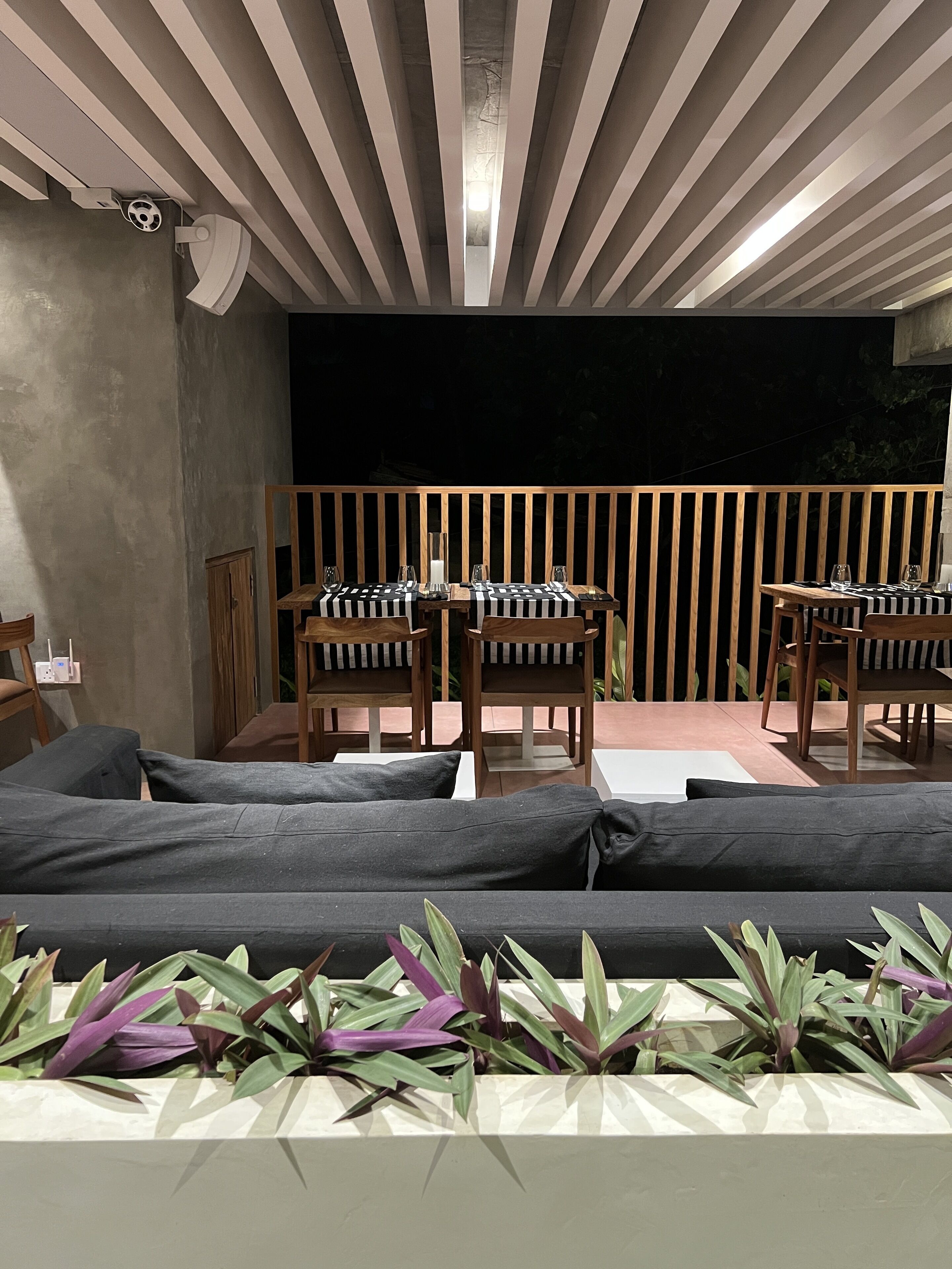 A modern outdoor seating area with wooden tables and chairs, a sofa, and a planter with green plants.