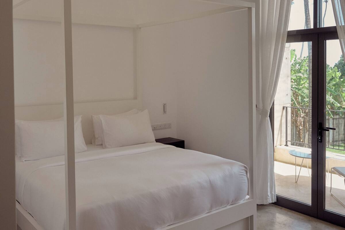 A white four-poster bed with white bedding, a ceiling fan, and a view of the outdoors through a glass door.