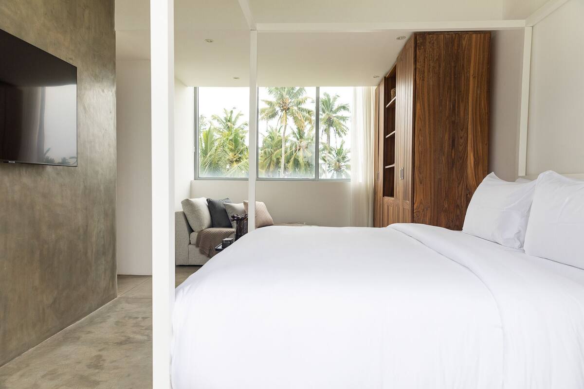 A modern bedroom with a large bed, a TV mounted on the wall, a wooden wardrobe, and a window with a view of palm trees.