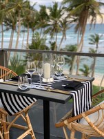 Mesa de comedor dispuesta con vasos, una vela y cubiertos, con vista a una playa con palmeras.