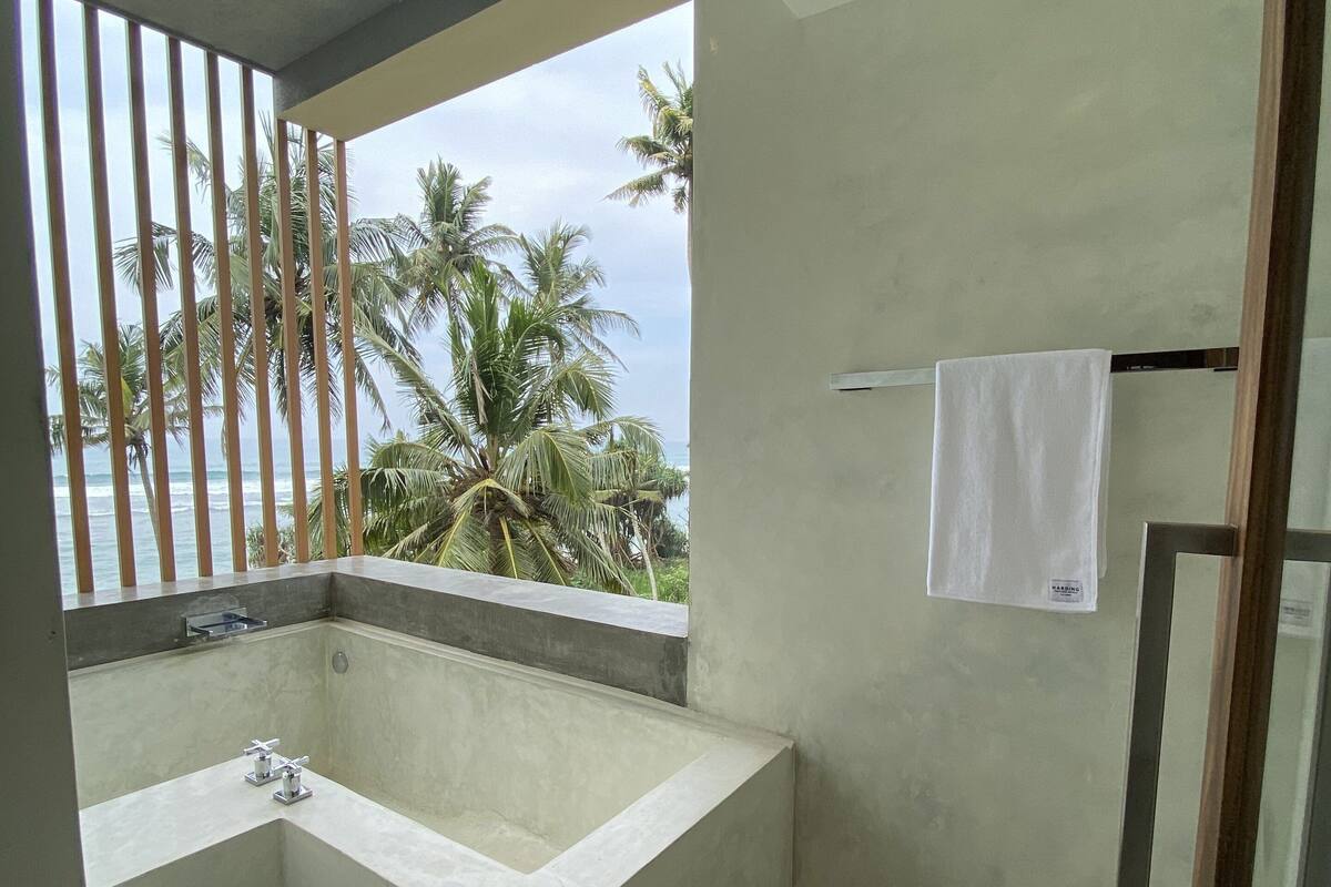 A bathroom with a large bathtub, a window with a view of palm trees and the sea, and a towel hanging on the wall.