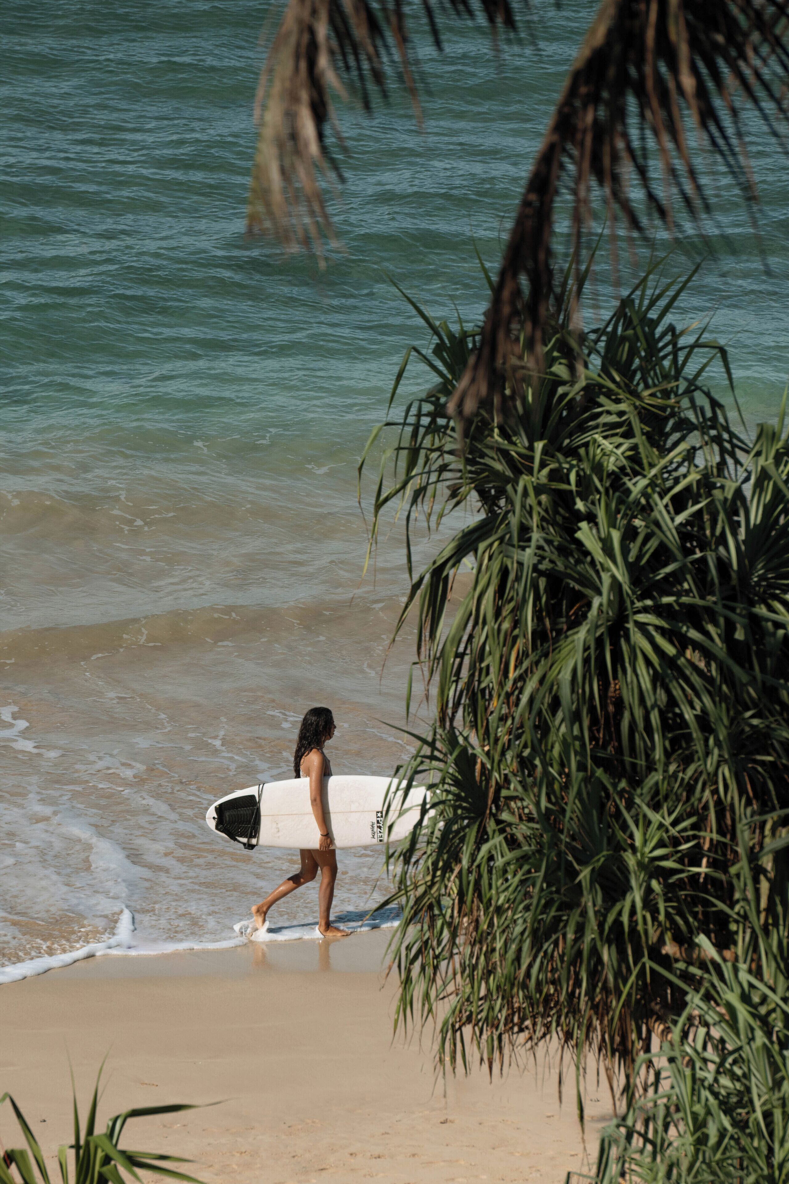 Una persona caminando sobre una playa arenosa con una tabla de surf, palmeras y olas del océano.