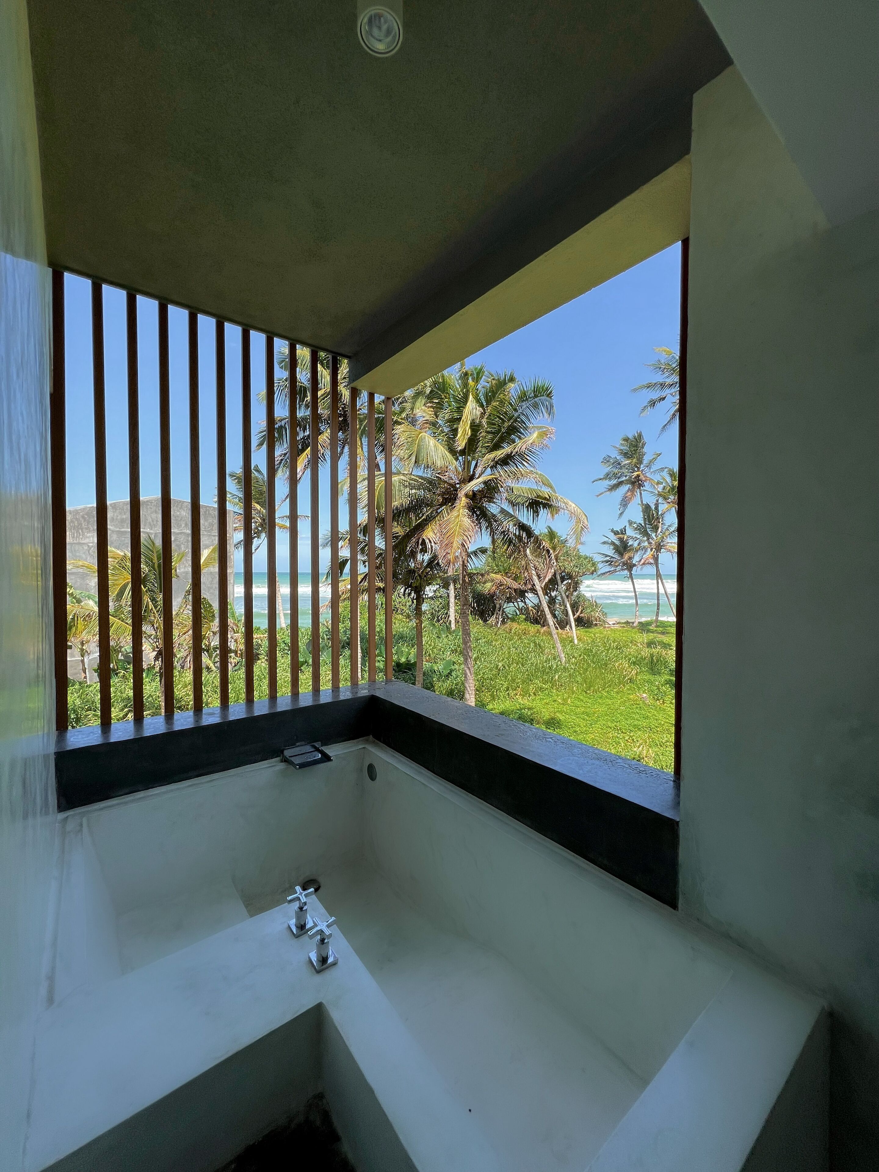 A bathroom with a bathtub, a window with a view of palm trees and the ocean, and a wooden railing.