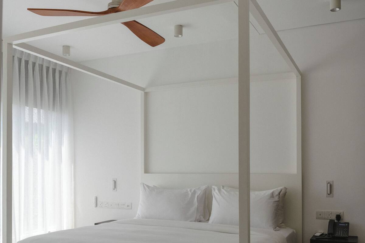 A white four-poster bed with a ceiling fan, a bedside table, and a nightstand.