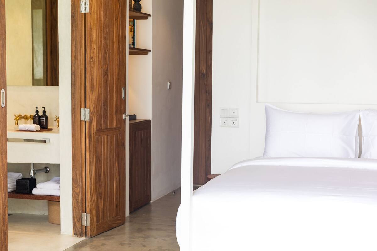 A hotel room with a wooden wardrobe, a bathroom with a mirror, and a bed with white bedding.