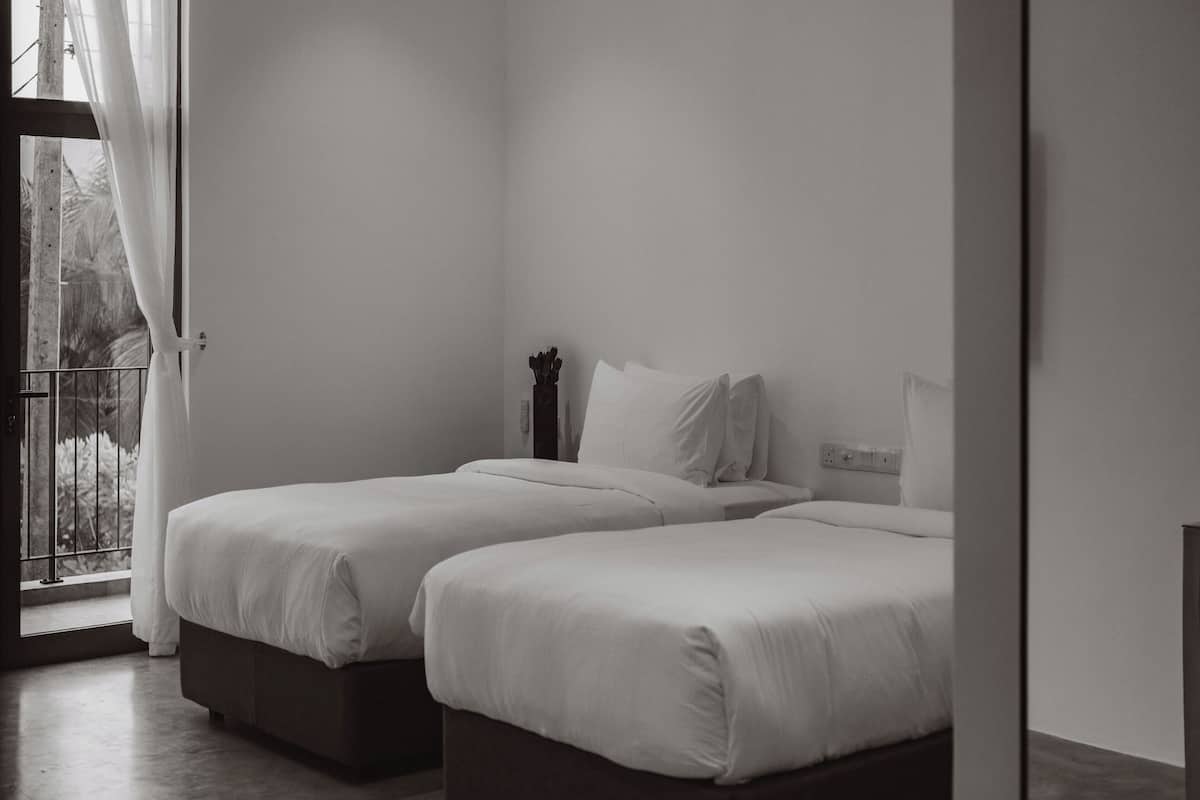 A minimalist bedroom with two beds, a window with sheer curtains, and a balcony view.