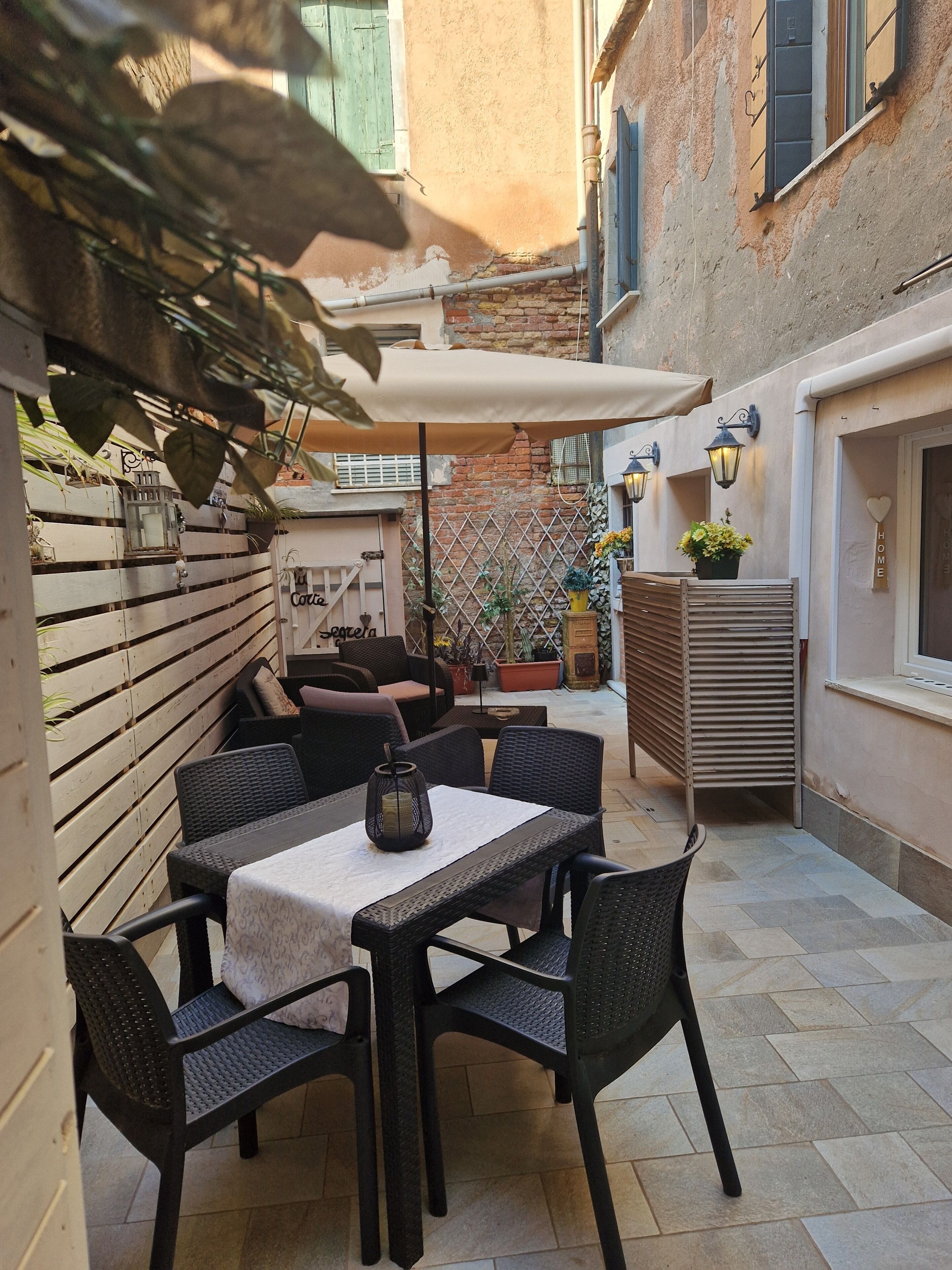 Eine Terrasse im Freien mit Tisch und Stühlen, einem Holzzaun, Topfpflanzen und einem an der Wand befestigten Laternenaufsatz.