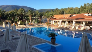 A pool area with lounge chairs and umbrellas, surrounded by palm trees and a mountain in the background. - Steps from the Ionian Sea, literally 10m from barefoot bliss! (Karavomylos)