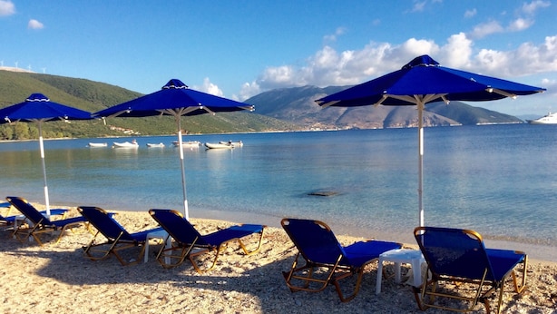 A beach with blue umbrellas and lounge chairs, a calm lake, boats, and mountains in the background. - Steps from the Ionian Sea, literally 10m from barefoot bliss! (Karavomylos)