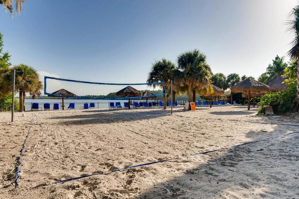 A beach volleyball court with net, blue lounge chairs, palm trees, and thatched-roof structures.