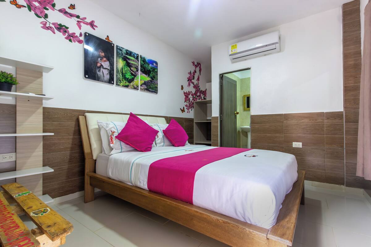 Un dormitorio con cama de madera, ropa de cama blanca y almohadas rosas. Hay decoraciones en la pared, un banco y una puerta que da a otra habitación.