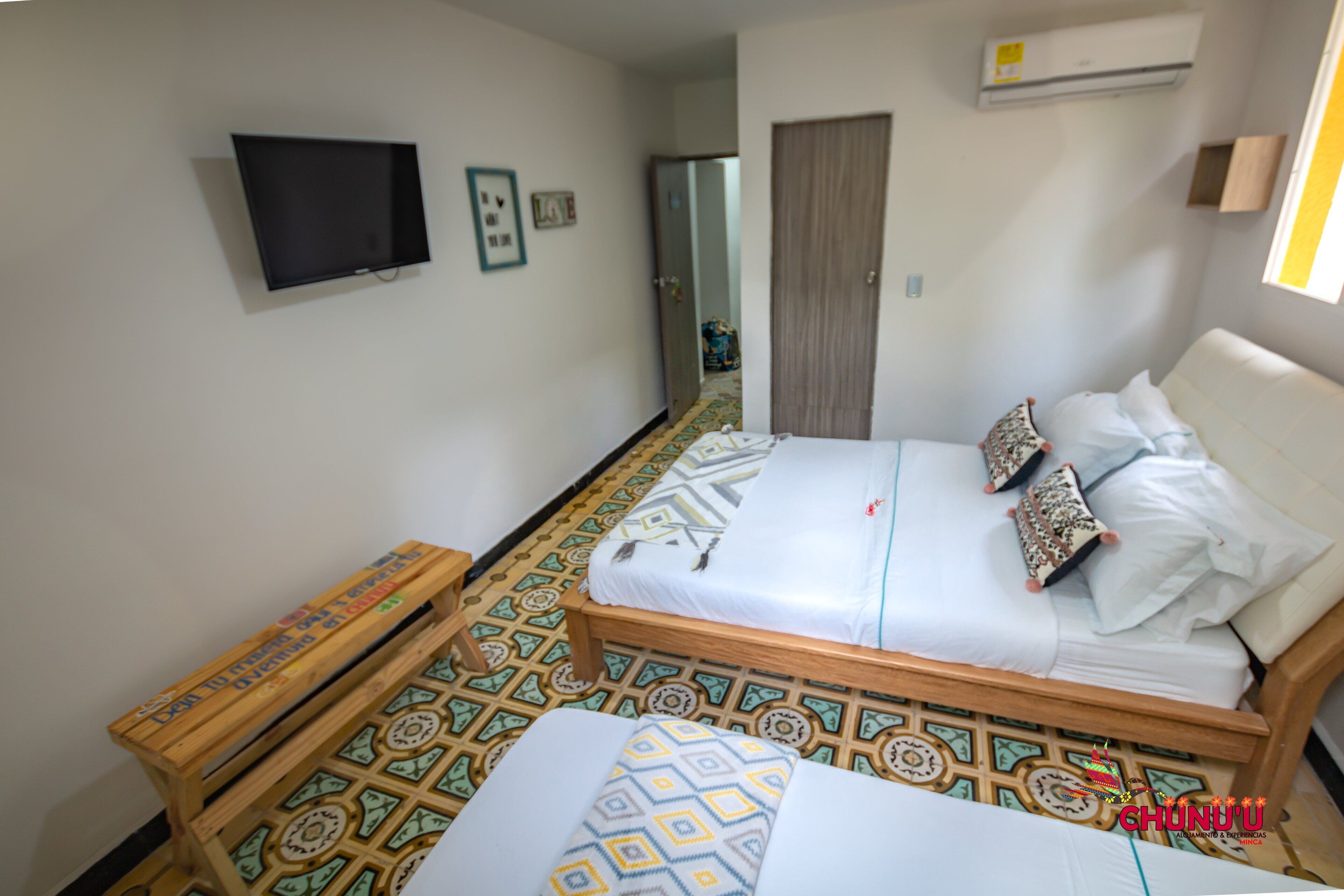 A bedroom with a bed, a TV mounted on the wall, a wooden bench, patterned floor tiles, and a door.