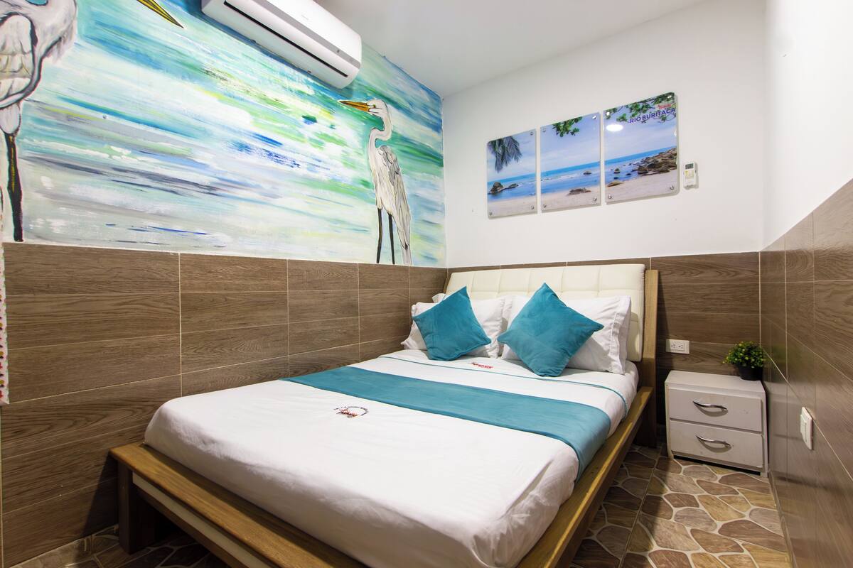 Una cama bien hecha con colcha azul y blanca, un cabecero de madera y una mesita de noche con una planta.