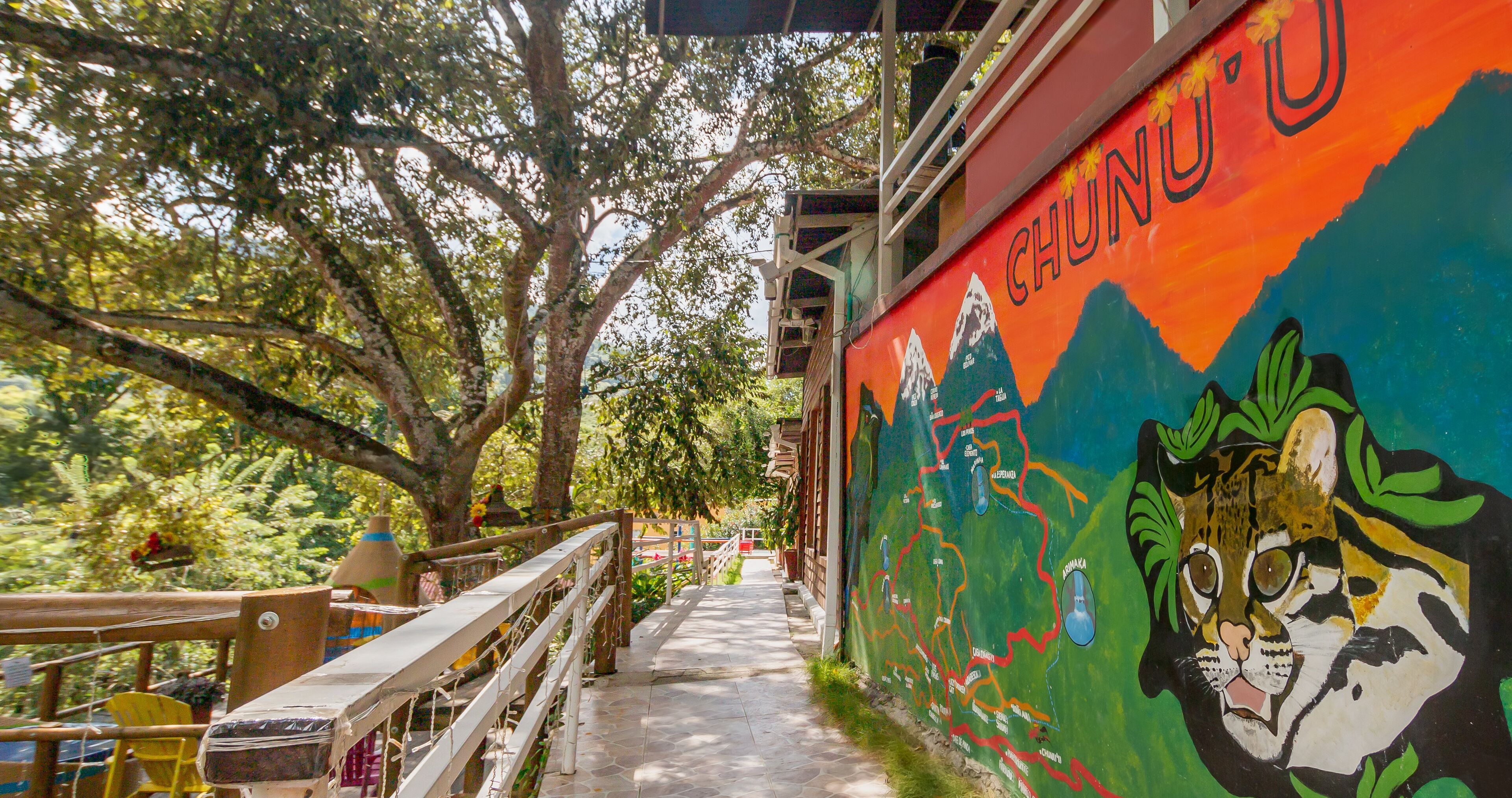 A pathway with a wooden railing, a mural with mountain and animal illustrations, and a view of trees and foliage.